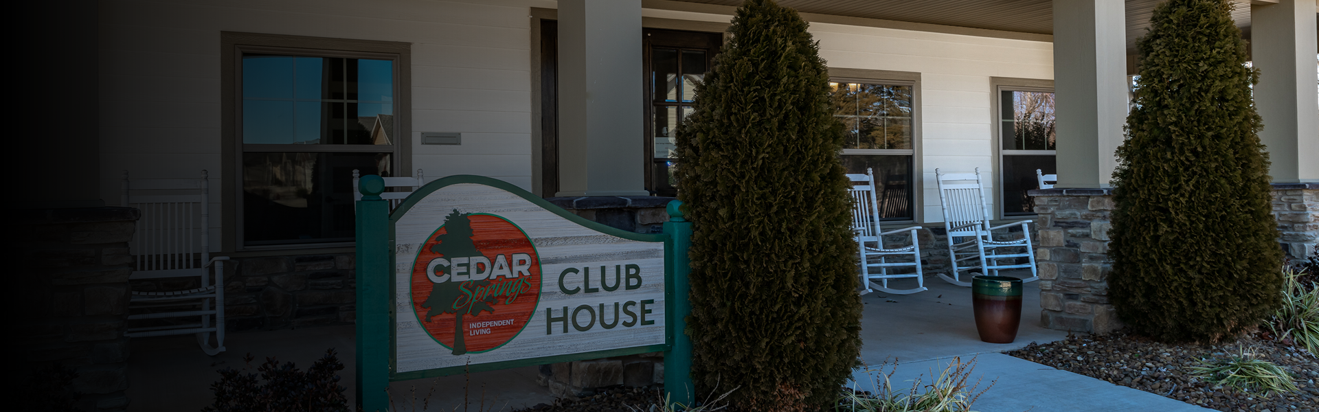 Front entrance of Cedar Springs Club House with a large sign on the porch and white rocking chairs nearby under two evergreens.