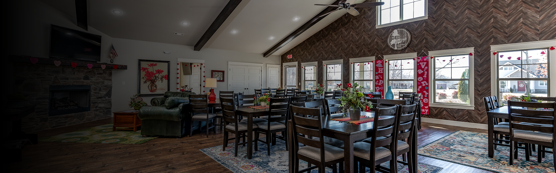 Communal dining room with dark wood tables and chairs, festive Valentine decorations, and large windows along a wood-paneled wall; stone fireplace on the left.