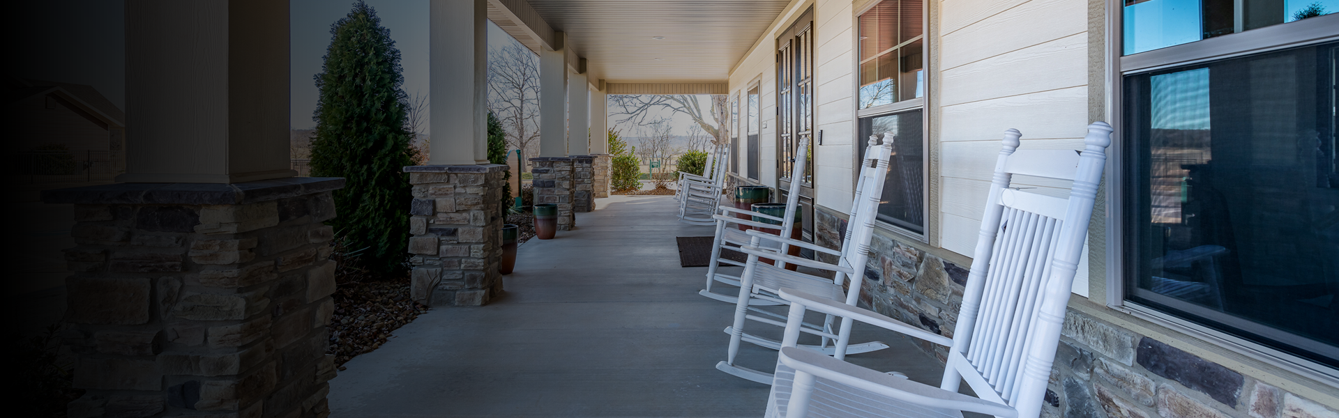 Row of white rocking chairs lined up along a covered front porch with stone columns and a beige house exterior