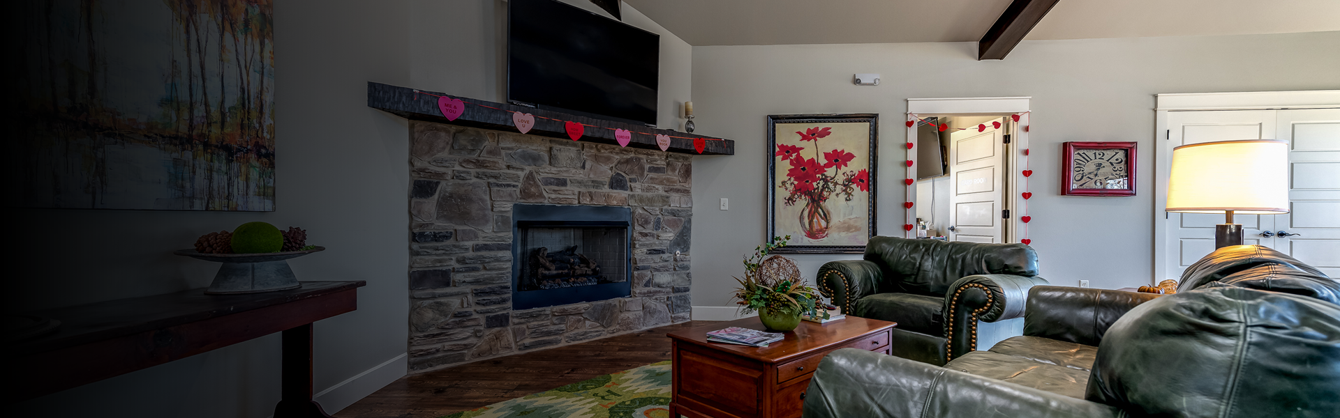 Cozy living room with a stone fireplace, heart garland, and dark green leather couches around a wooden coffee table.