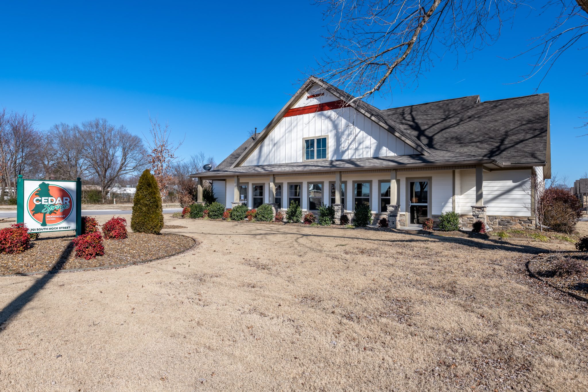 White, single-story building with a steep gabled roof and a front porch, surrounded by shrubs, under a bright blue sky; a Cedar Springs sign is in the foreground.