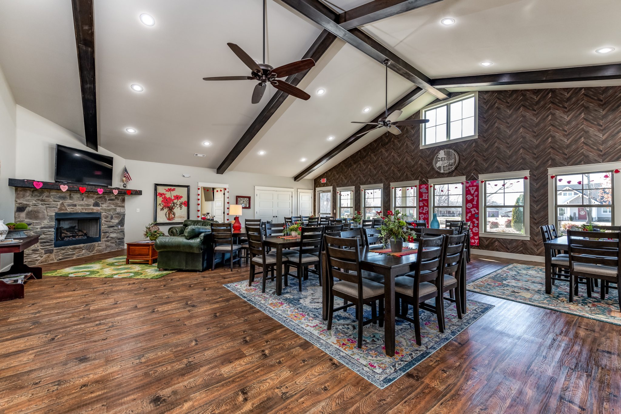 Rustic event hall with dark wood vaulted beams, ceiling fans, and rows of tables and chairs set for a gathering; Valentine decorations along windows and mantel near a stone fireplace.