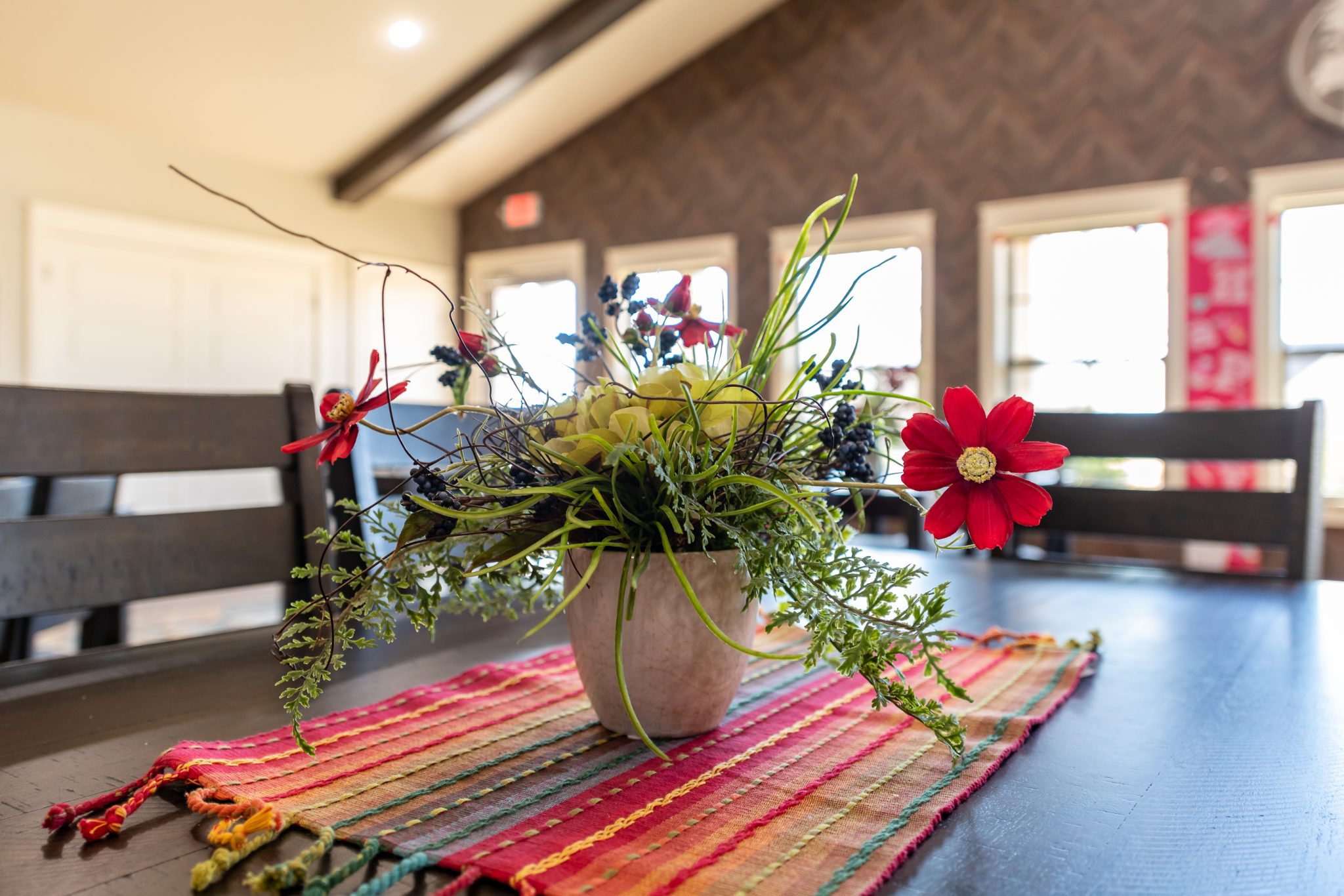 Close-up of a colorful floral arrangement in a beige pot on a dark dining table with a bright, striped runner indoors