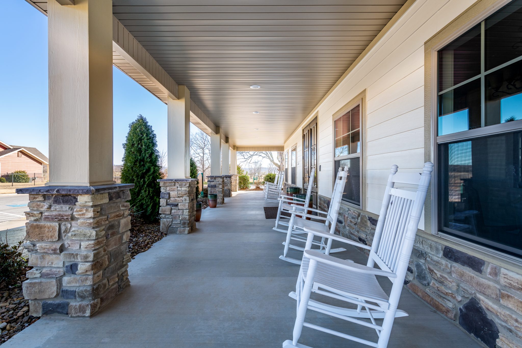 Covered porch with white rocking chairs along the house and stone columns beneath a overhang.