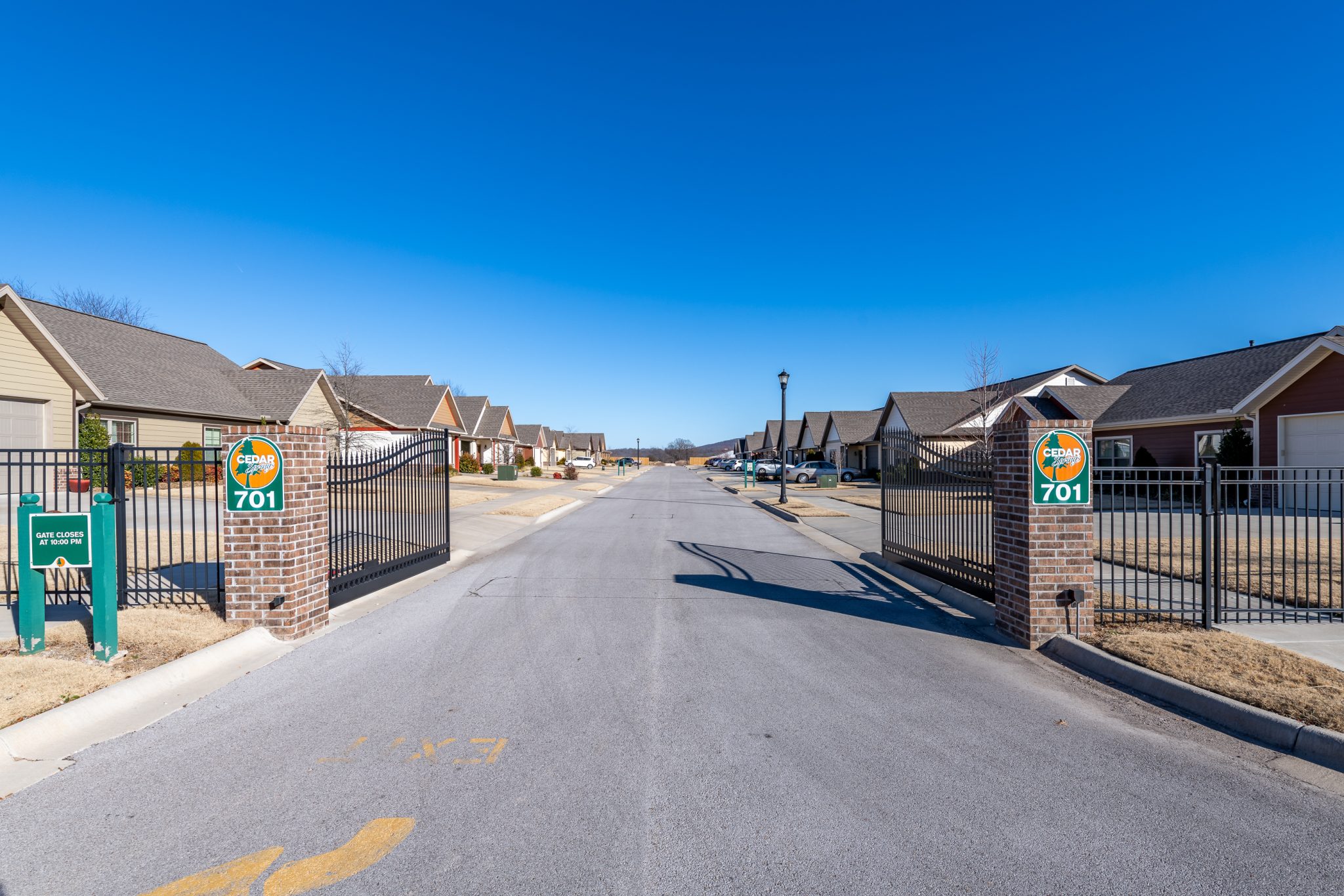 Gated entrance to a suburban neighborhood with brick pillars and signs reading Cedar Springs 701, row of houses along a sunny street under a blue sky.