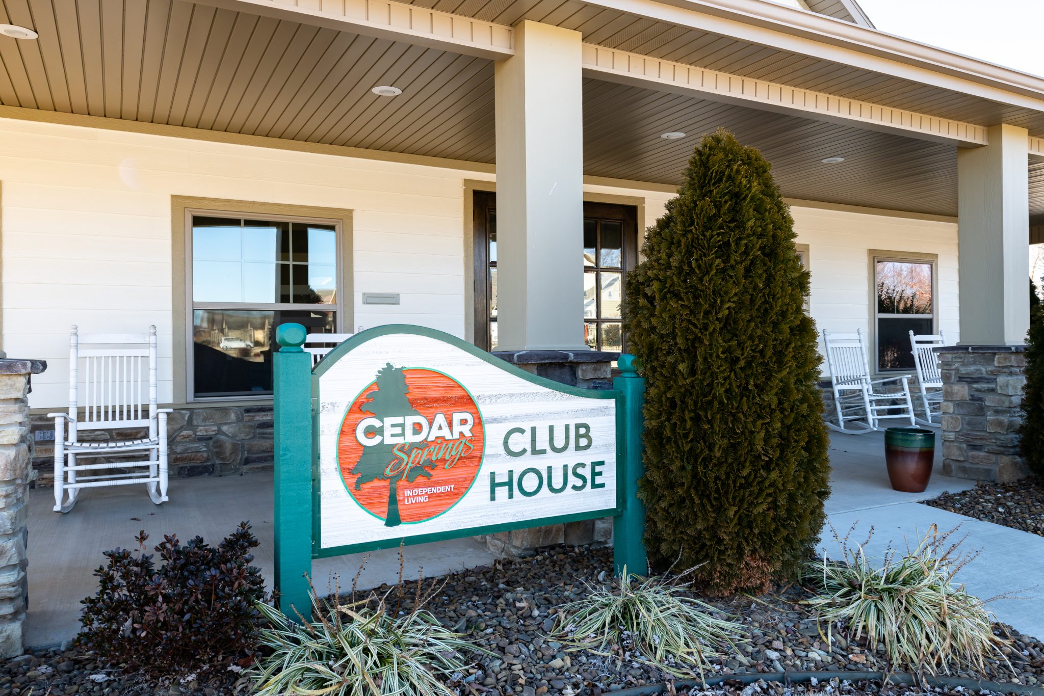Front view of Cedar Springs Club House with a colorful sign, white rocking chairs on the porch, and a beige siding building behind it.